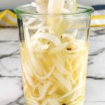 Pickled white onions in a glass jar with peppercorn brine and sliced onions lifted with chopsticks.