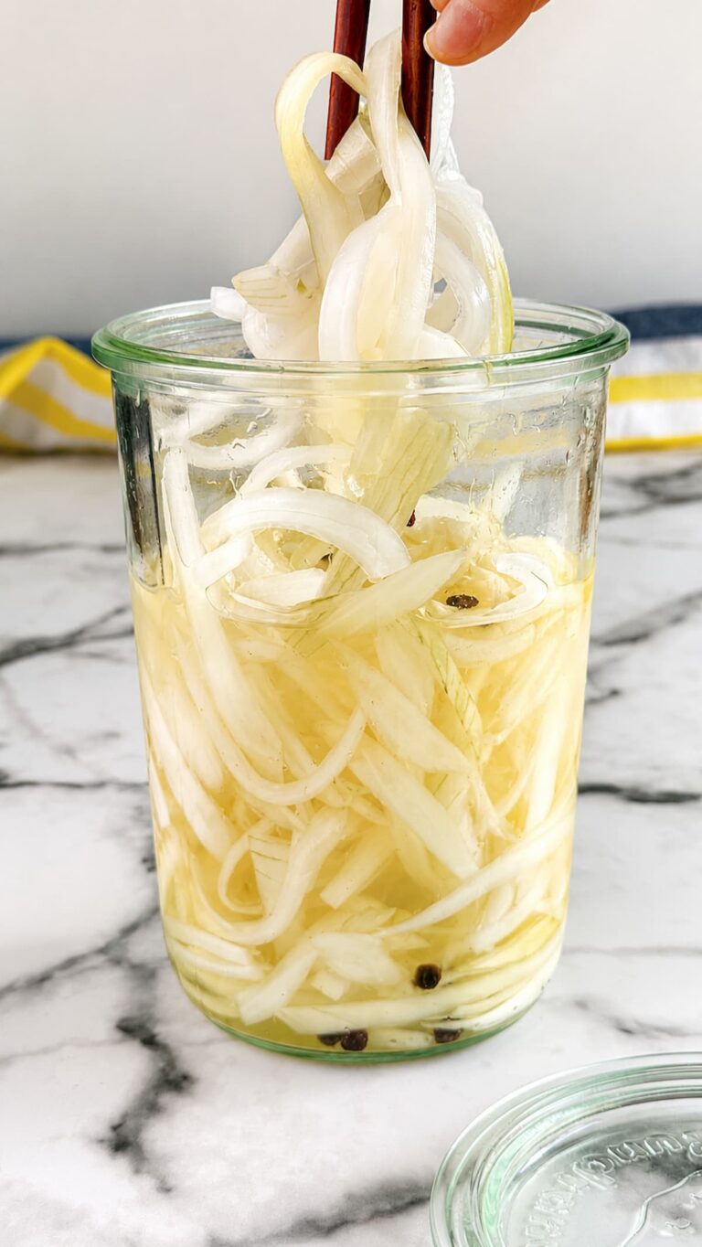 Pickled white onions in a glass jar with peppercorn brine and sliced onions lifted with chopsticks.