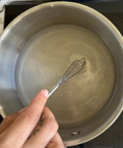 hand with a little whisk whisking sugar in water in a stainless steel saucepan on a burner.