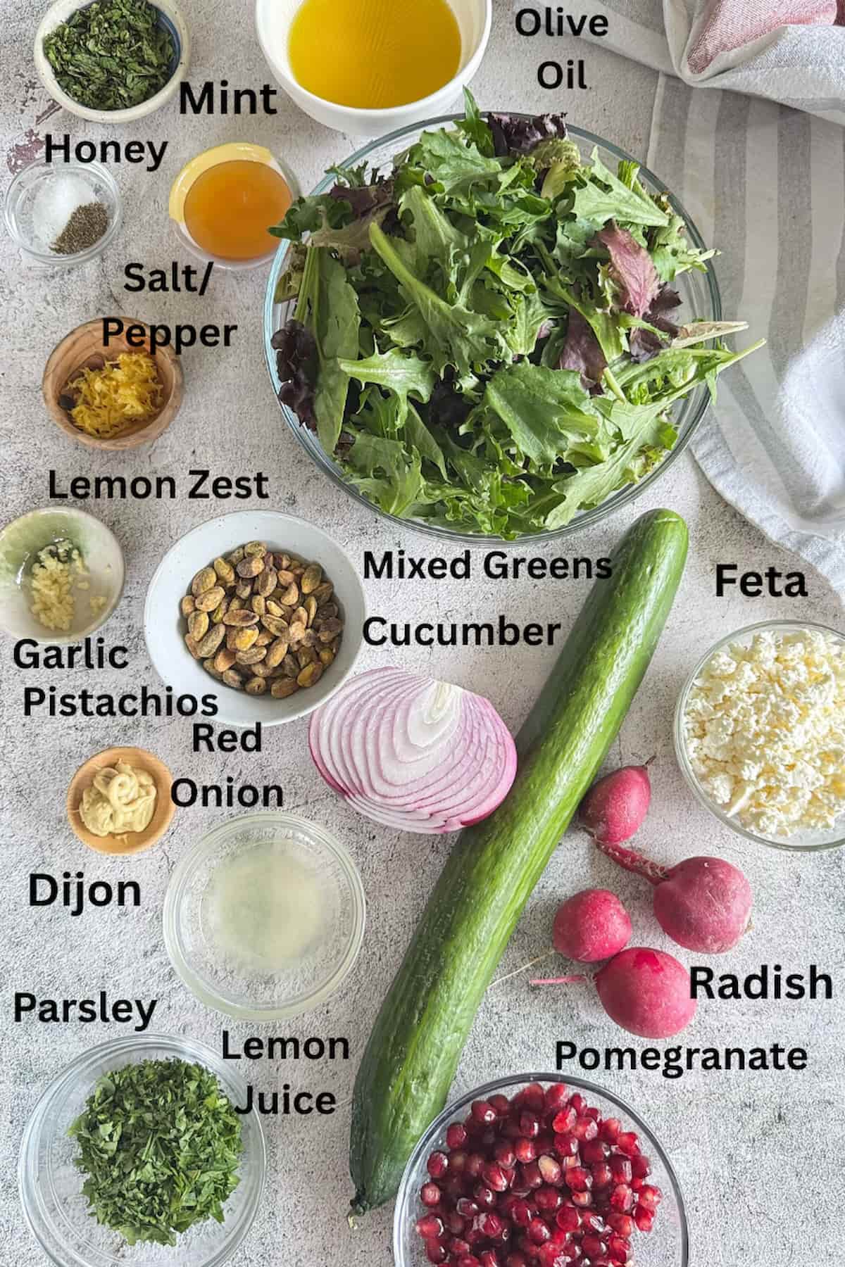 ingredients for pomegranate and feta salad laid out next to each other in different bowls - mint, honey, olive oil, salt, pepper, lemon juice, lemon zest, dijon, garlic, pistachios, feta, pomegranate, parsley, mixed greens, radish, red onion, and cucumber.
