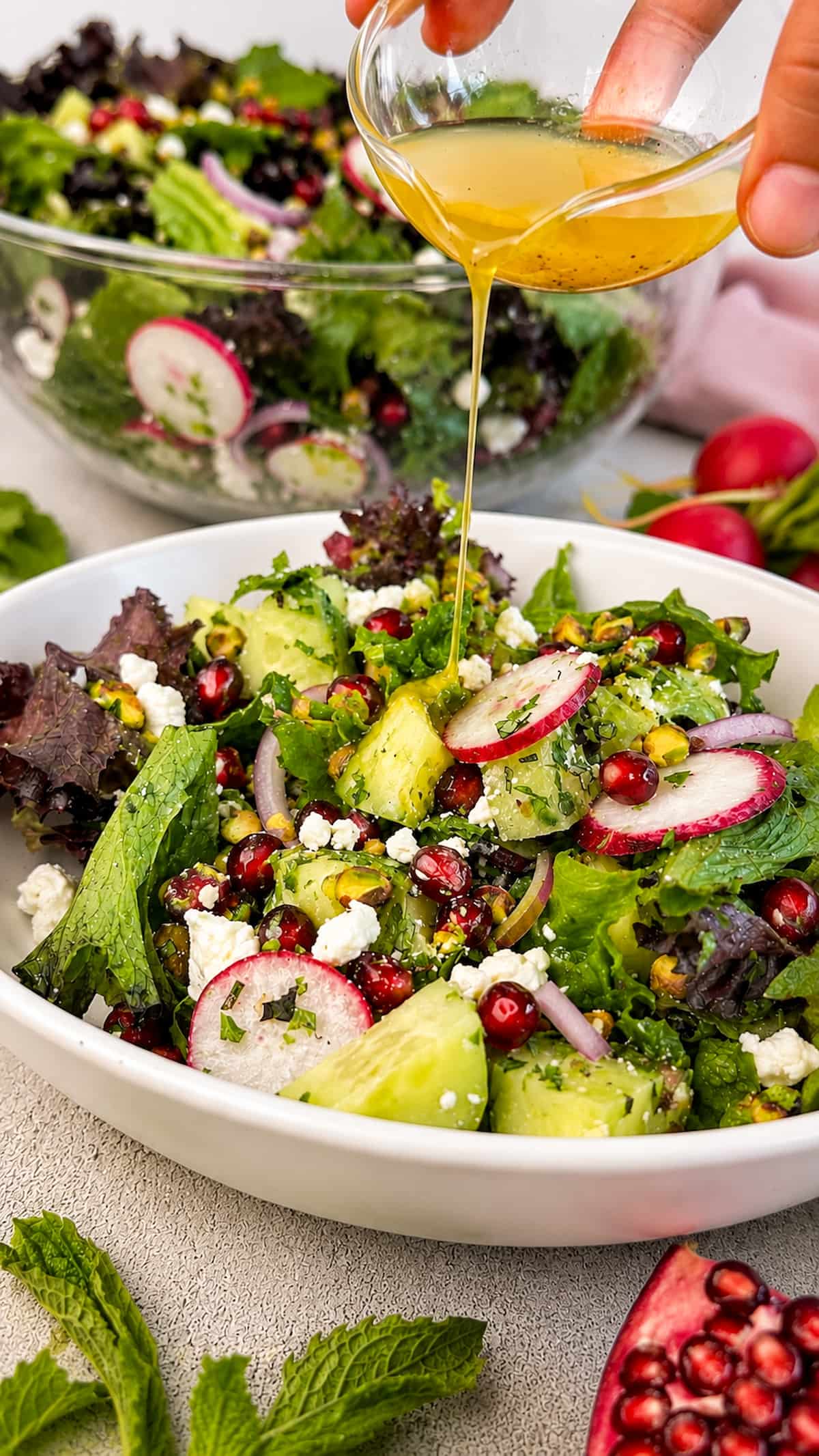 Close up of a fresh pomegranate feta salad with mixed greens, cucumber, radishes, red onion, pistachios, crumbled feta, and mint as a homemade lemon vinaigrette is drizzled over the top.
