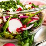 Close up side view of a fresh pomegranate feta salad with mixed greens, radishes, cucumber, red onion, pistachios, fresh herbs, and crumbled feta in a clear glass serving bowl.