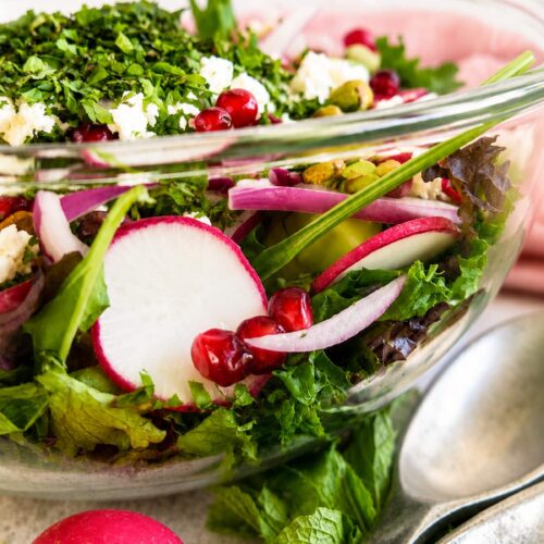Close up side view of a fresh pomegranate feta salad with mixed greens, radishes, cucumber, red onion, pistachios, fresh herbs, and crumbled feta in a clear glass serving bowl.