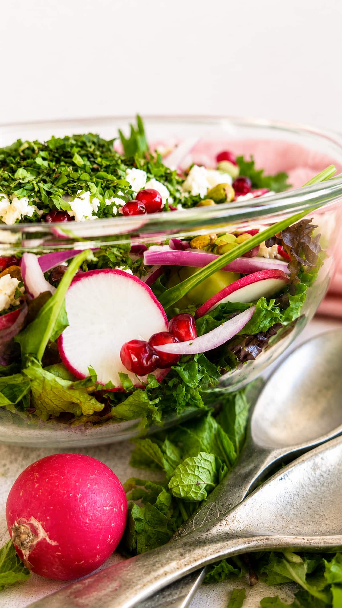 Close up side view of a fresh pomegranate feta salad with mixed greens, radishes, cucumber, red onion, pistachios, fresh herbs, and crumbled feta in a clear glass serving bowl.