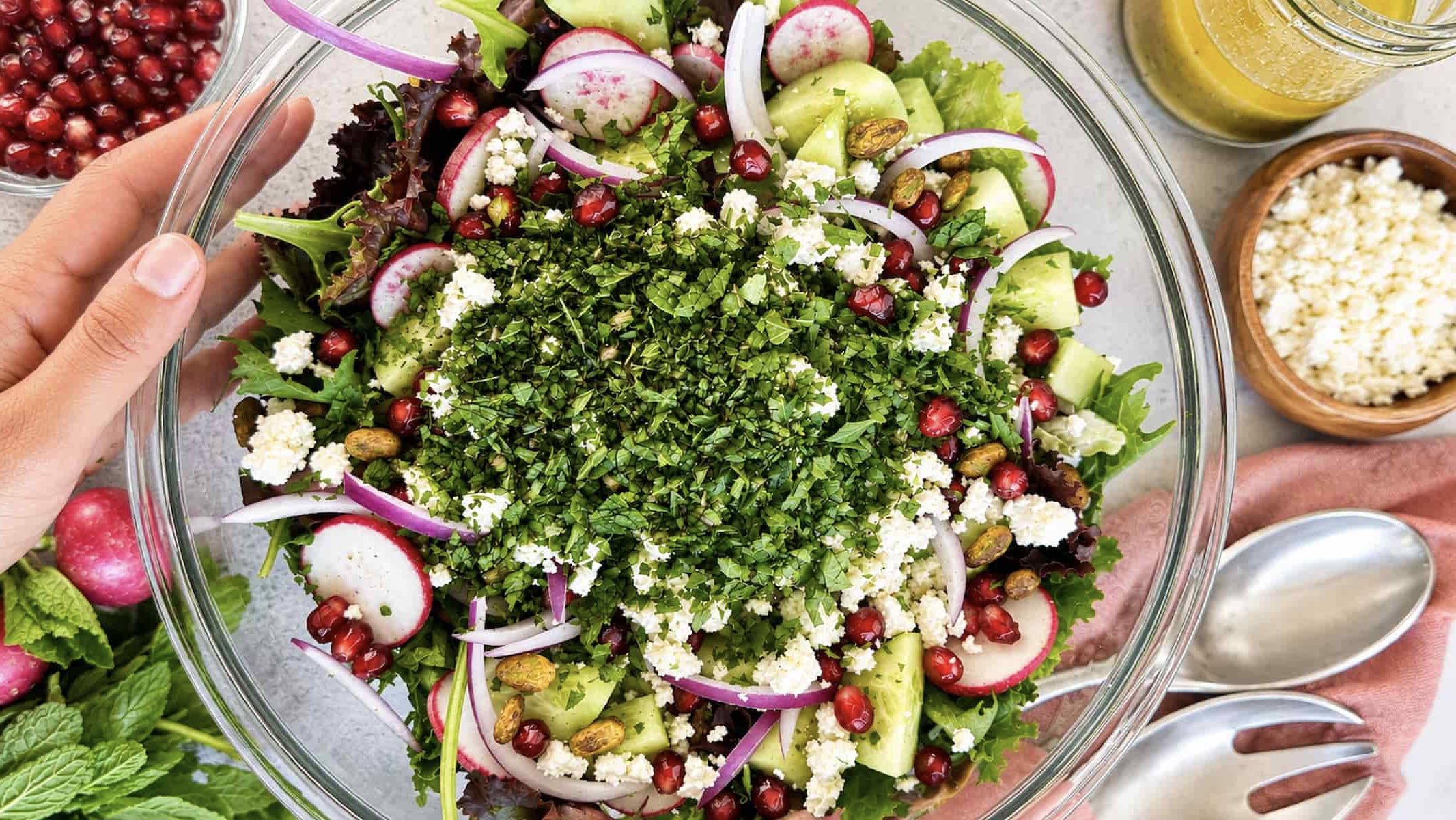 Fresh pomegranate feta salad with mixed greens, cucumber, radishes, red onion, pistachios, fresh herbs, and a lemon vinaigrette in a large glass serving bowl.