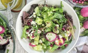 Overhead view of a fresh pomegranate feta salad with mixed greens, cucumber, radishes, red onion, crumbled feta, and pomegranate arils tossed in a lemon vinaigrette and served in a white bowl.