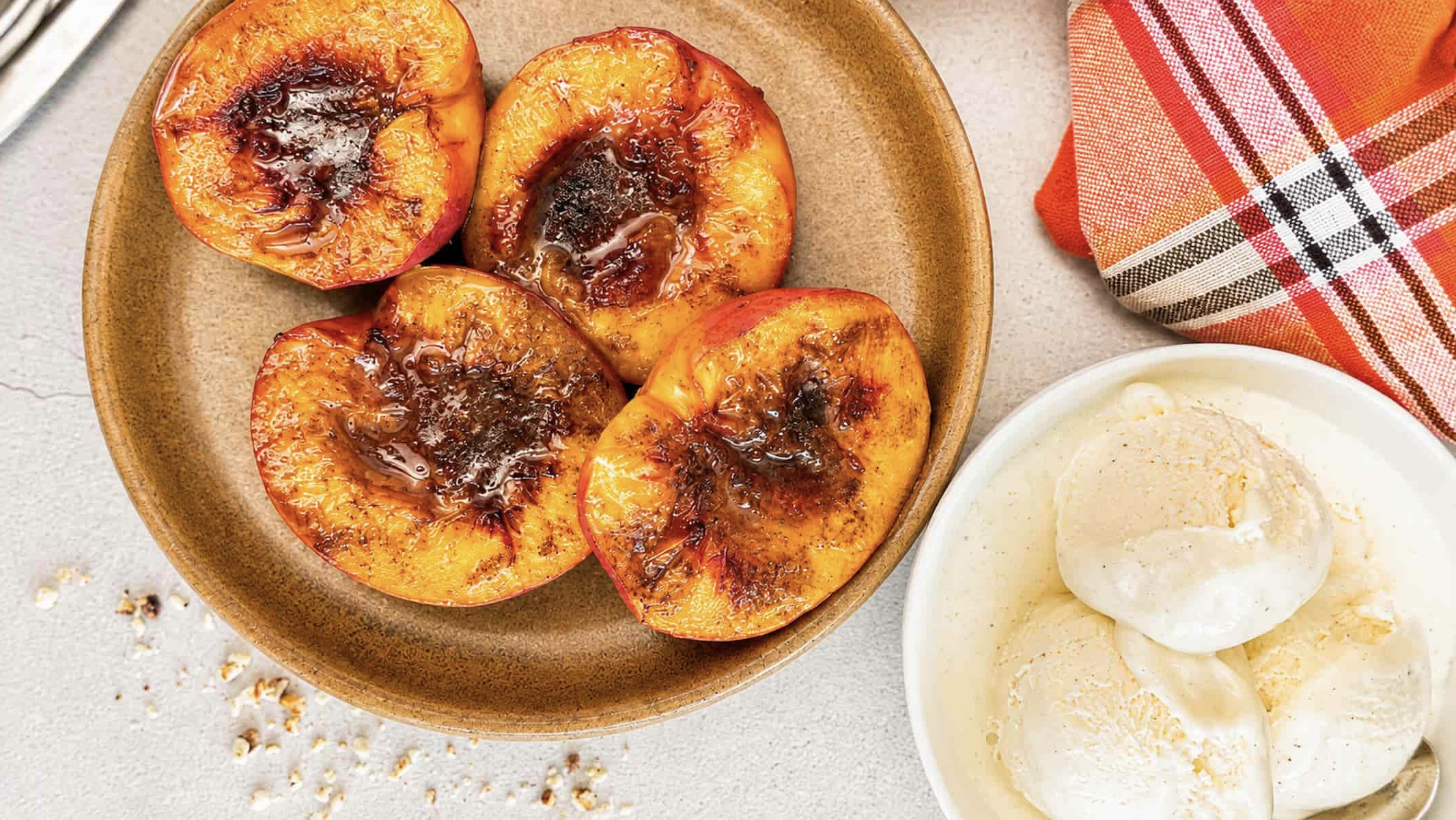 4 Air fryer peach halves caramelized with cinnamon and brown sugar on a plate, next to a bowl with 3 scoops of vanilla ice cream.