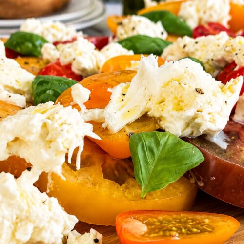 Close up of Burrata Caprese salad with creamy burrata, heirloom tomatoes, cherry tomatoes, fresh basil, and olive oil on a wooden serving board.