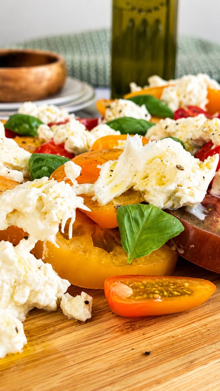 Close up of Burrata Caprese salad with creamy burrata, heirloom tomatoes, cherry tomatoes, fresh basil, and olive oil on a wooden serving board.