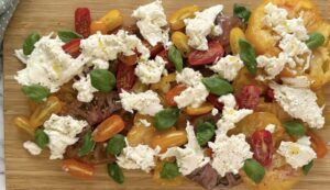 burrata caprese salad with mixed coloured heirloom and cherry tomatoes, torn burrata, fresh basil, and salt and pepper on a wooden board.