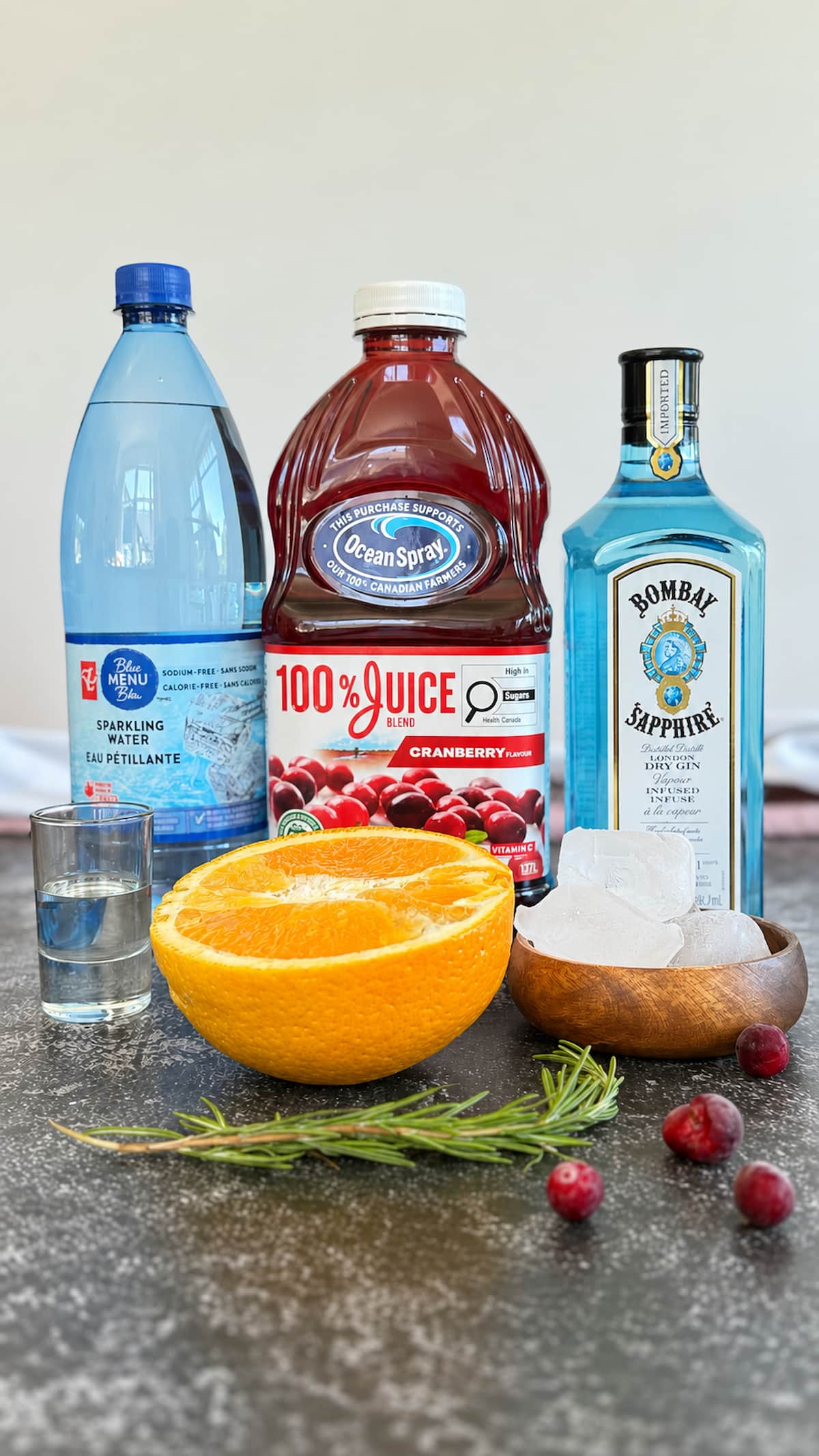 Ingredients for a cranberry gin fizz including gin, cranberry juice, sparkling water, orange, rosemary, ice, and a shot of gin.