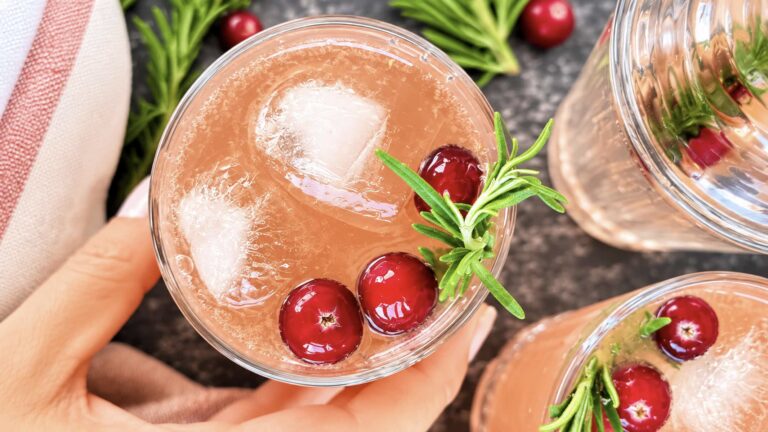 Hand holding a cranberry gin fizz garnished with fresh cranberries, rosemary, and ice.