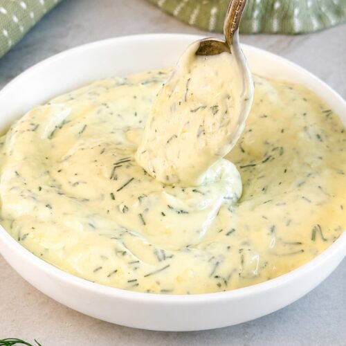 Creamy dill mayonnaise in a white bowl with fresh chopped dill and a spoon lifting a dollop.