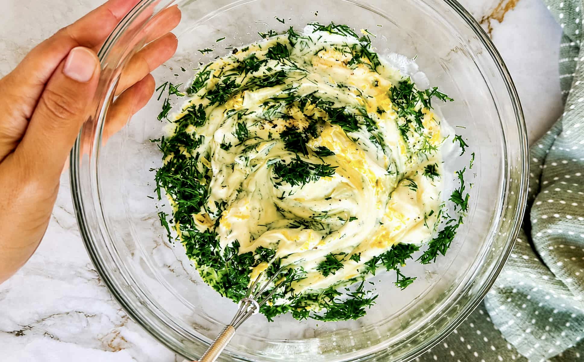 Dill mayonnaise being mixed with fresh dill, lemon zest, and seasonings in a glass bowl.