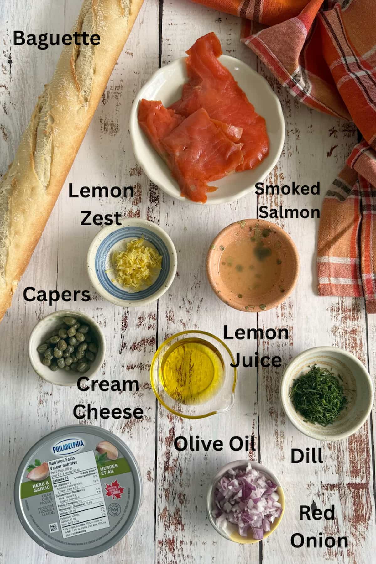 ingredients for smoked salmon crostini laid out in separate bowls - capers, lemon zest, lemon juice, smoked salmon, dill, red onion, olive oil, cream cheese, and a baguette.