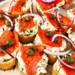Hand reaching for smoked salmon crostini topped with herbed cream cheese, red onion, capers, and fresh dill.