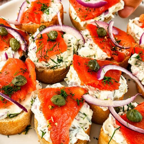 Hand reaching for smoked salmon crostini topped with herbed cream cheese, red onion, capers, and fresh dill.