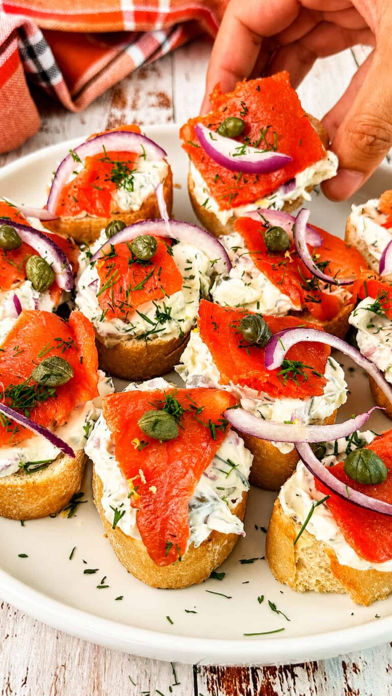 Hand reaching for smoked salmon crostini topped with herbed cream cheese, red onion, capers, and fresh dill.