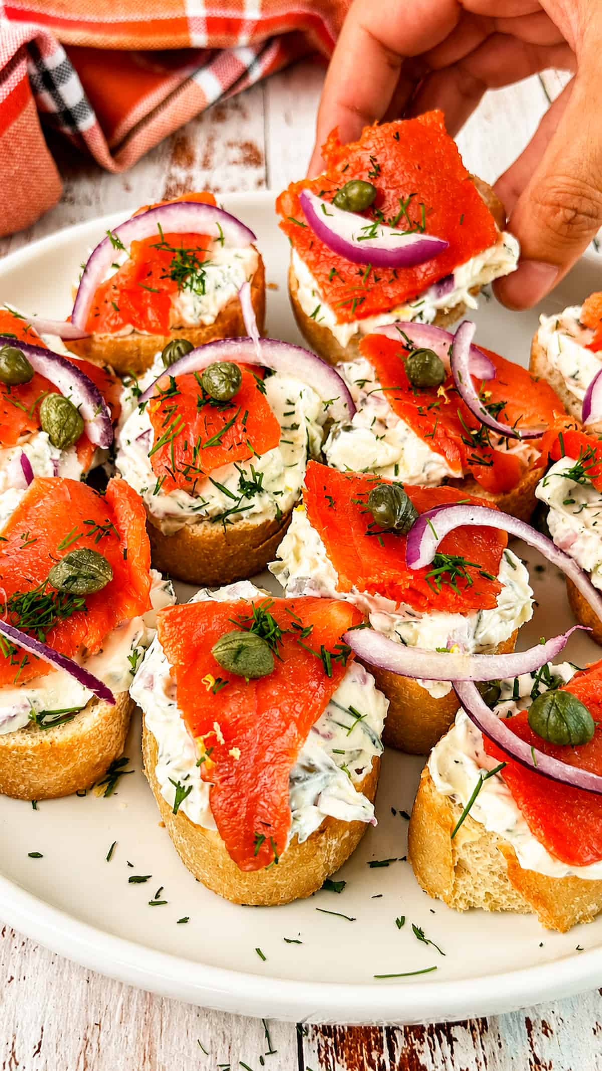 Hand reaching for smoked salmon crostini topped with herbed cream cheese, red onion, capers, and fresh dill.