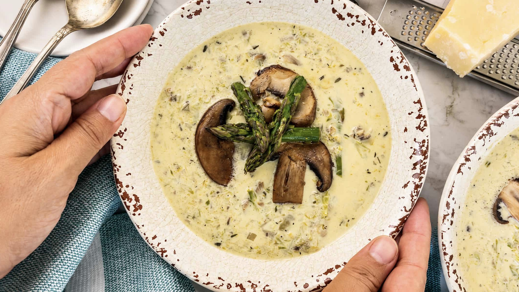 Creamy mushroom and asparagus soup topped with sautéed mushrooms and roasted asparagus, served in a rustic bowl held by two hands.