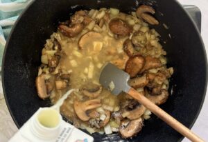 broth being poured into a pot of browned mushrooms and onions, small spatula in the pot.
