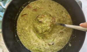 chunky green soup with asparagus and mushrooms in a black dutch oven pot, hand holding a ladle in the pot.