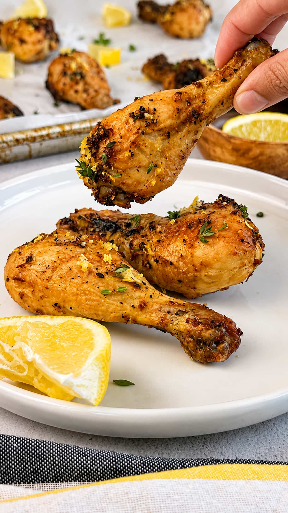 Lemon pepper chicken drumsticks on a plate with fresh thyme, lemon zest, and lemon wedges, hand grabbing one.