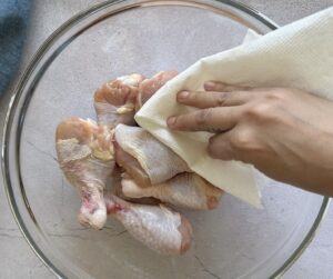 hand with a piece of paper towel patting raw chicken drumsticks dry in a large glass bowl.