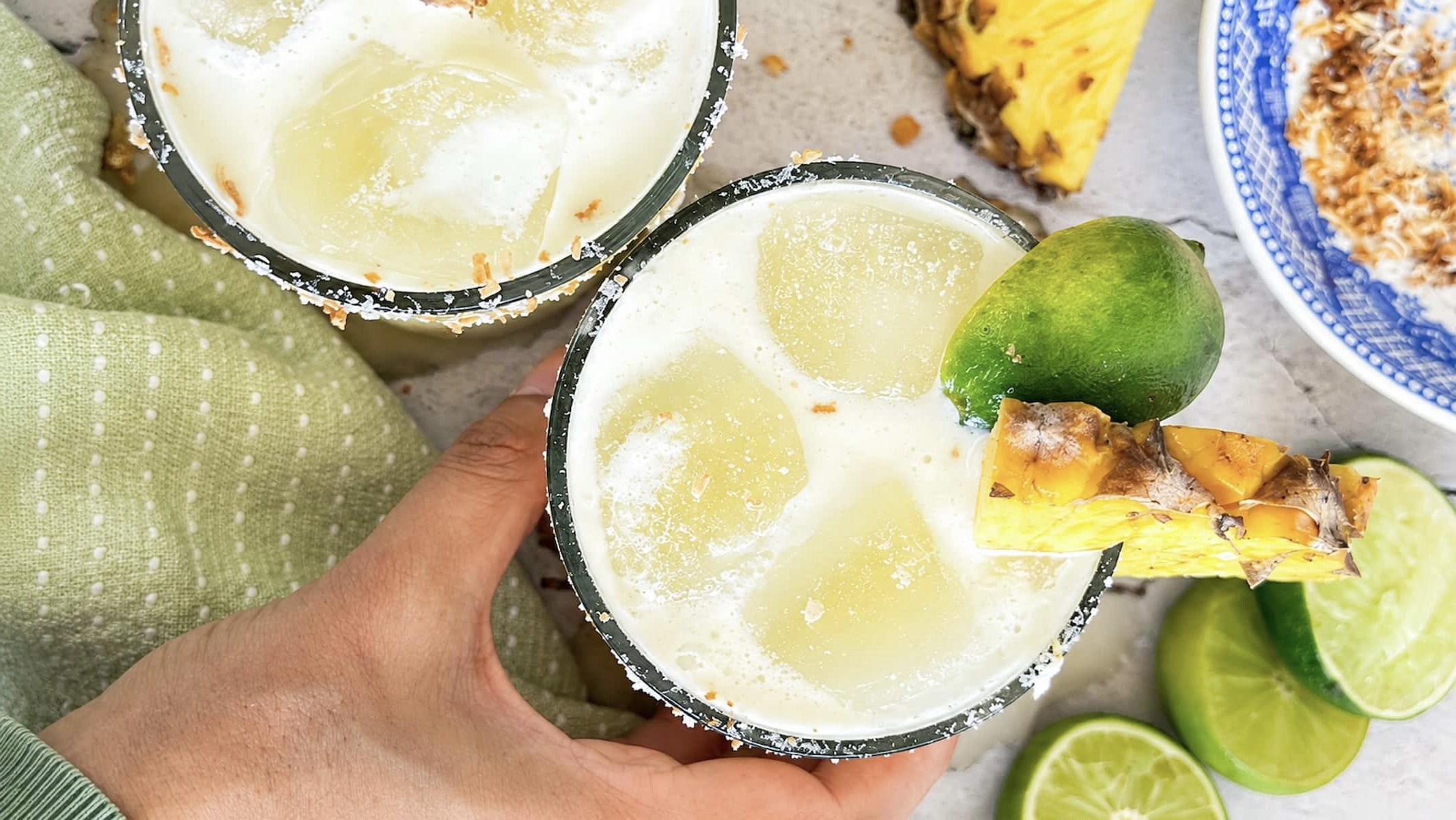 Hand holding a pineapple coconut margarita with fresh pineapple, lime, ice, and a toasted coconut salt rim.