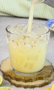 Pineapple coconut margarita being poured over ice with fresh pineapple, lime, and a toasted coconut salt rim.