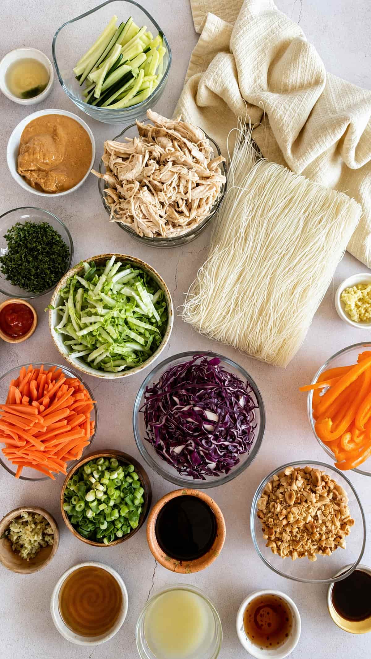 ingredients for thai chicken noodle salad laid out in separate bowls - rice noodles, shredded chicken, cucumber, peanut butter, napa cabbage, red cabbage, bell pepper, carrots, peanuts, green onions, cilantro, mint, garlic, sriracha, lime juice, fish sauce, sesame oil, honey, soy sauce, ginger, and water.