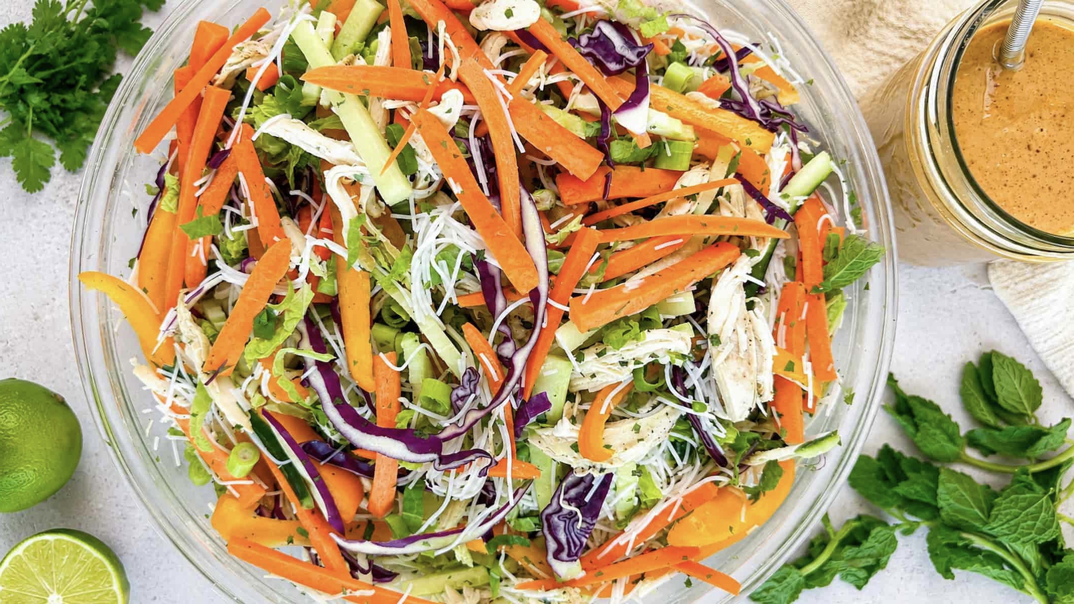 Thai chicken noodle salad with rice noodles, shredded chicken, crisp vegetables, fresh herbs, and creamy peanut dressing before tossing.