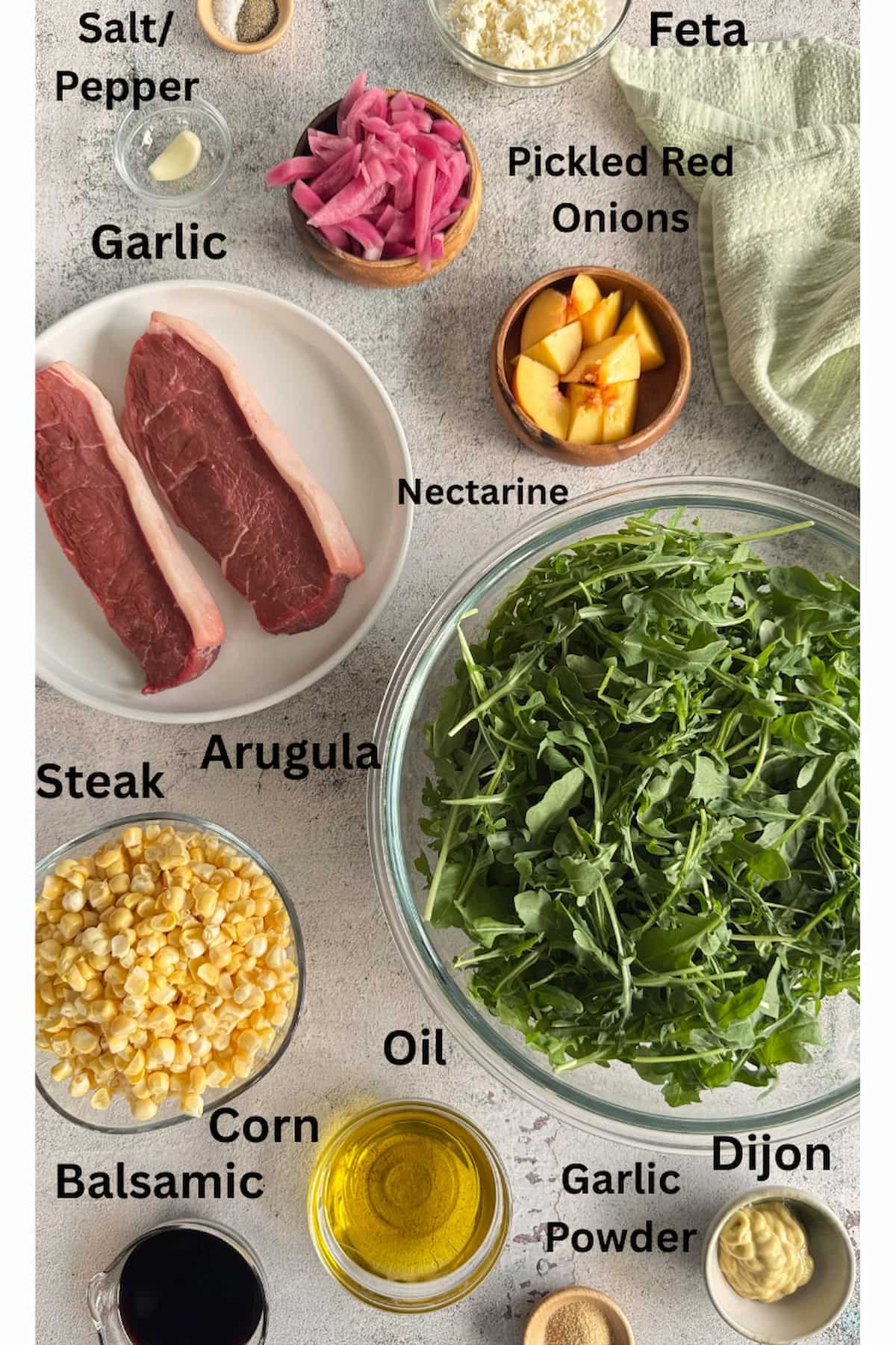 ingredients for arugula salad with steak laid out in separate bowls - arugula, steak, salt, pepper, garlic powder, oil, feta, pickled red onions, corn, nectarine, dijon, balsamic, and garlic.
