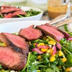 Arugula steak salad topped with juicy sliced steak, sweet corn, pickled red onions, crumbled cheese, and a creamy homemade dressing.