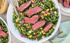 Arugula steak salad topped with sliced medium-rare steak, sweet corn, pickled red onions, crumbled feta, and a creamy homemade dressing.