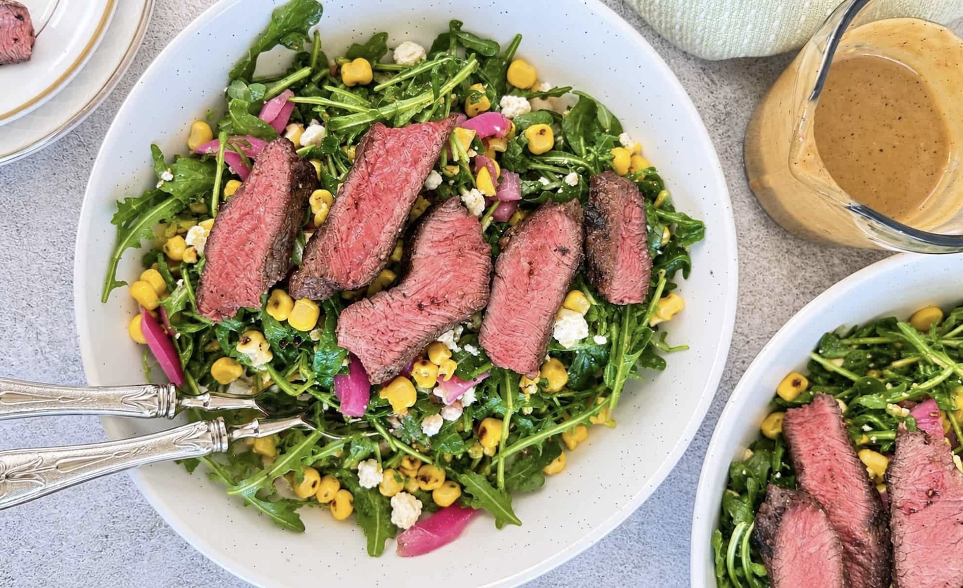 Arugula steak salad with tender medium-rare steak, charred corn, pickled red onions, crumbled feta, and creamy homemade dressing served on the side.