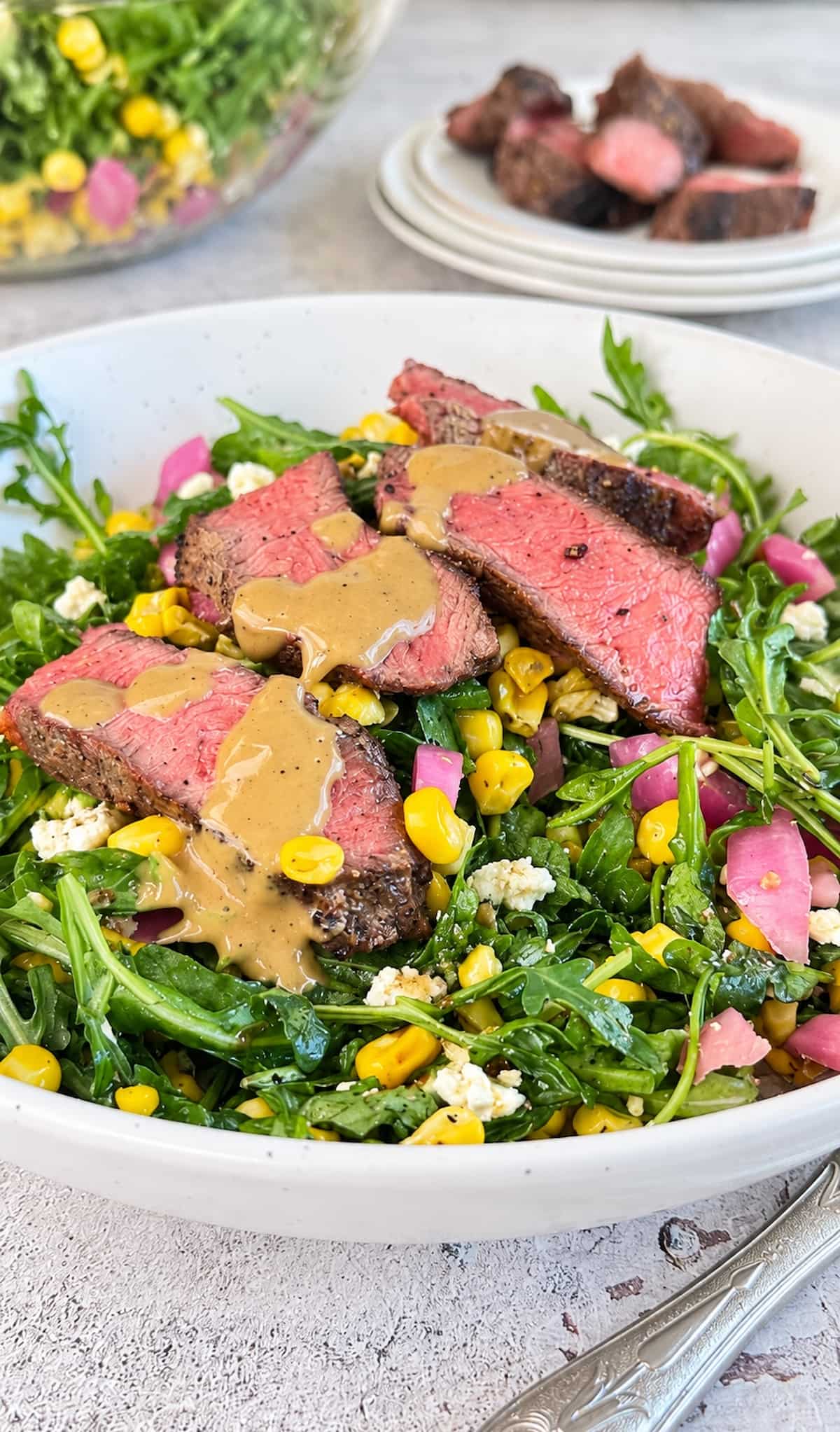 Arugula steak salad with juicy sliced steak, sweet corn, pickled red onions, crumbled feta, and creamy dressing drizzled over top.