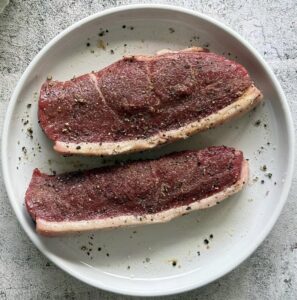 two raw steaks seasoned with salt and pepper on a white plate.
