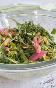 arugula tossed with balsamic, pickled red onions, corn, and feta cheese in a large glass bowl.