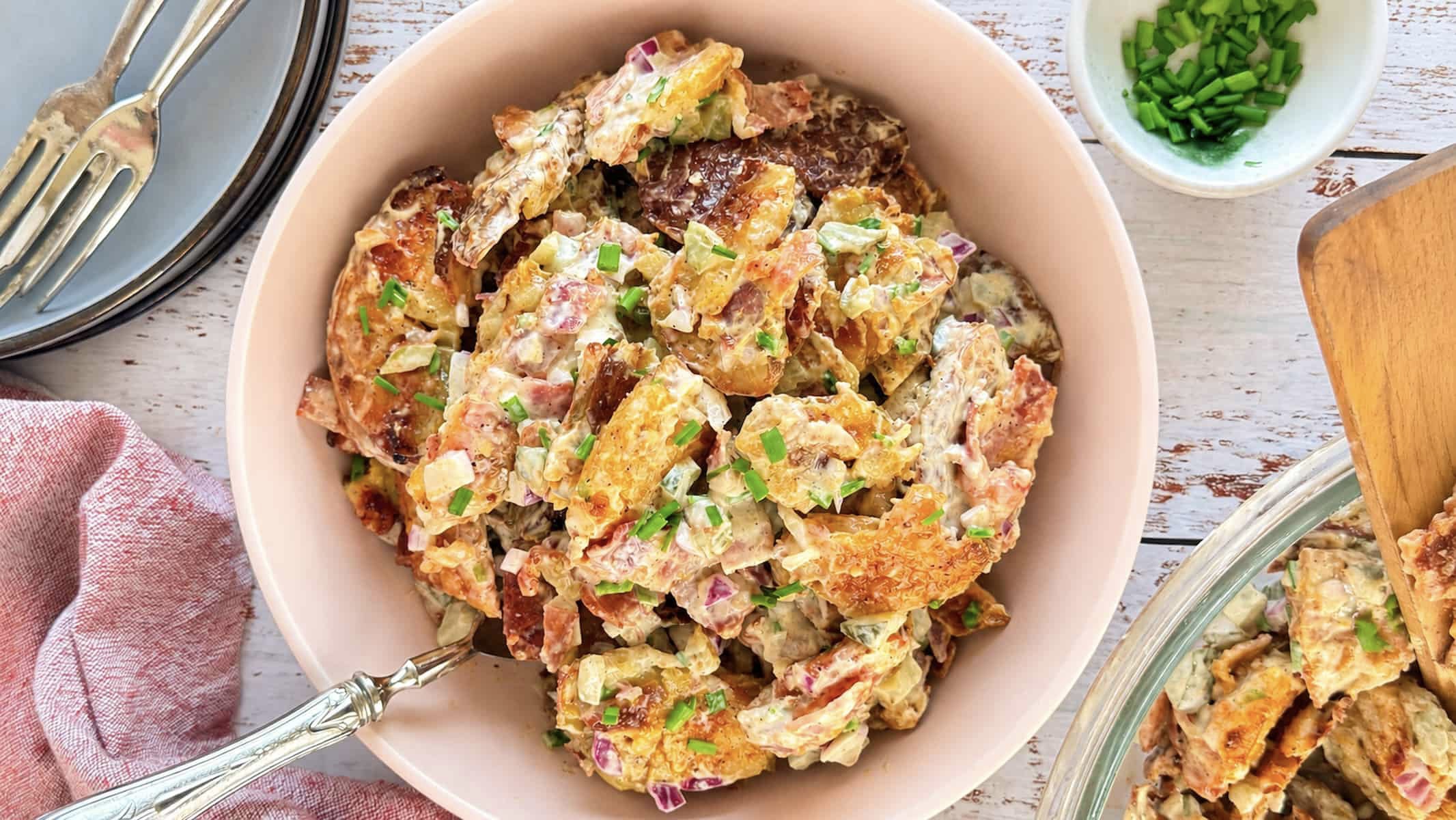 Smashed potato salad with crispy bacon, pickles, red onion, and fresh chives in a creamy dressing.