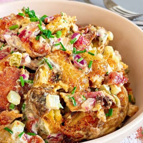 Smashed potato salad with crispy bacon, pickles, red onion, and fresh chives in a creamy dressing.