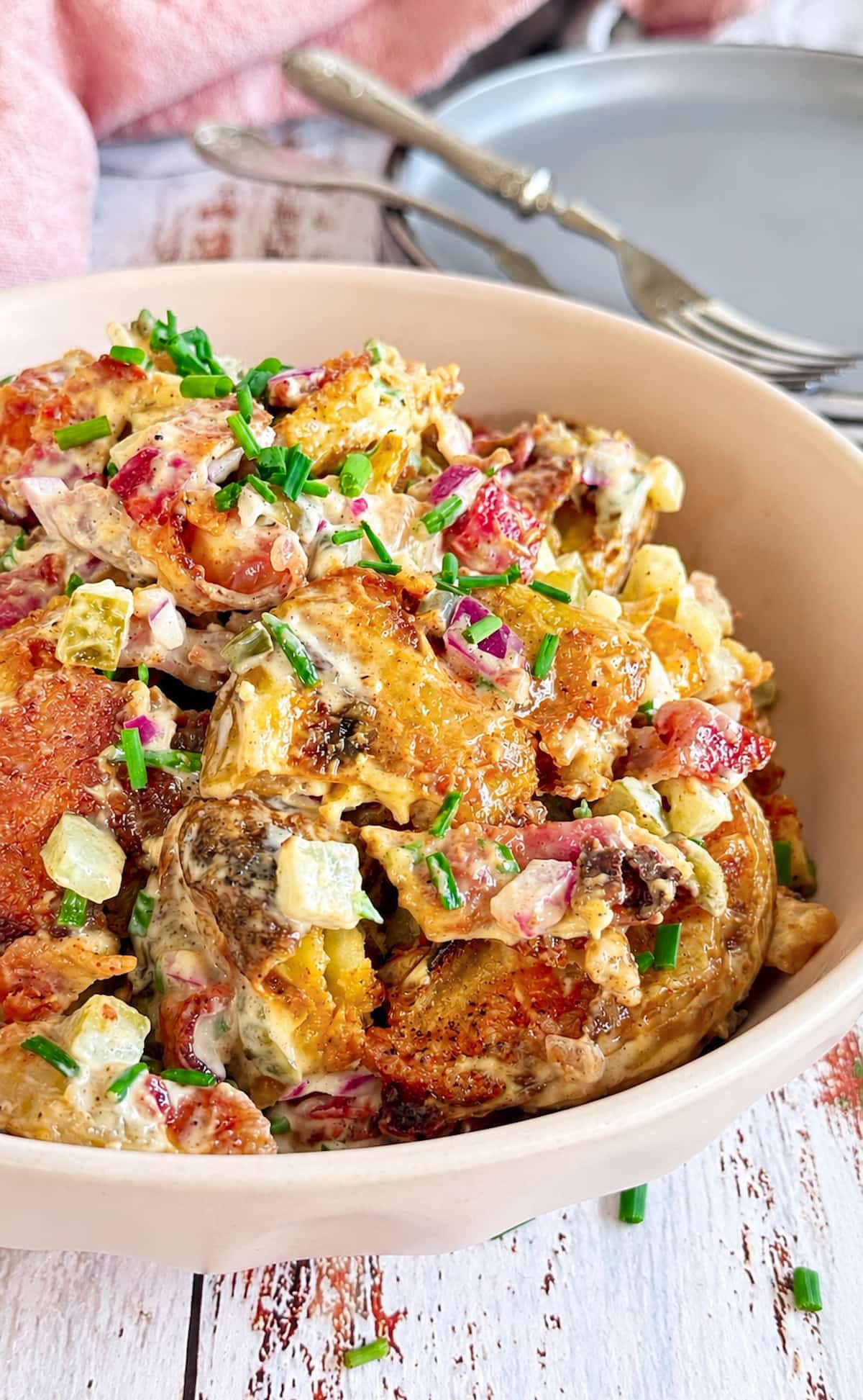 Smashed potato salad with crispy bacon, pickles, red onion, and fresh chives in a creamy dressing.