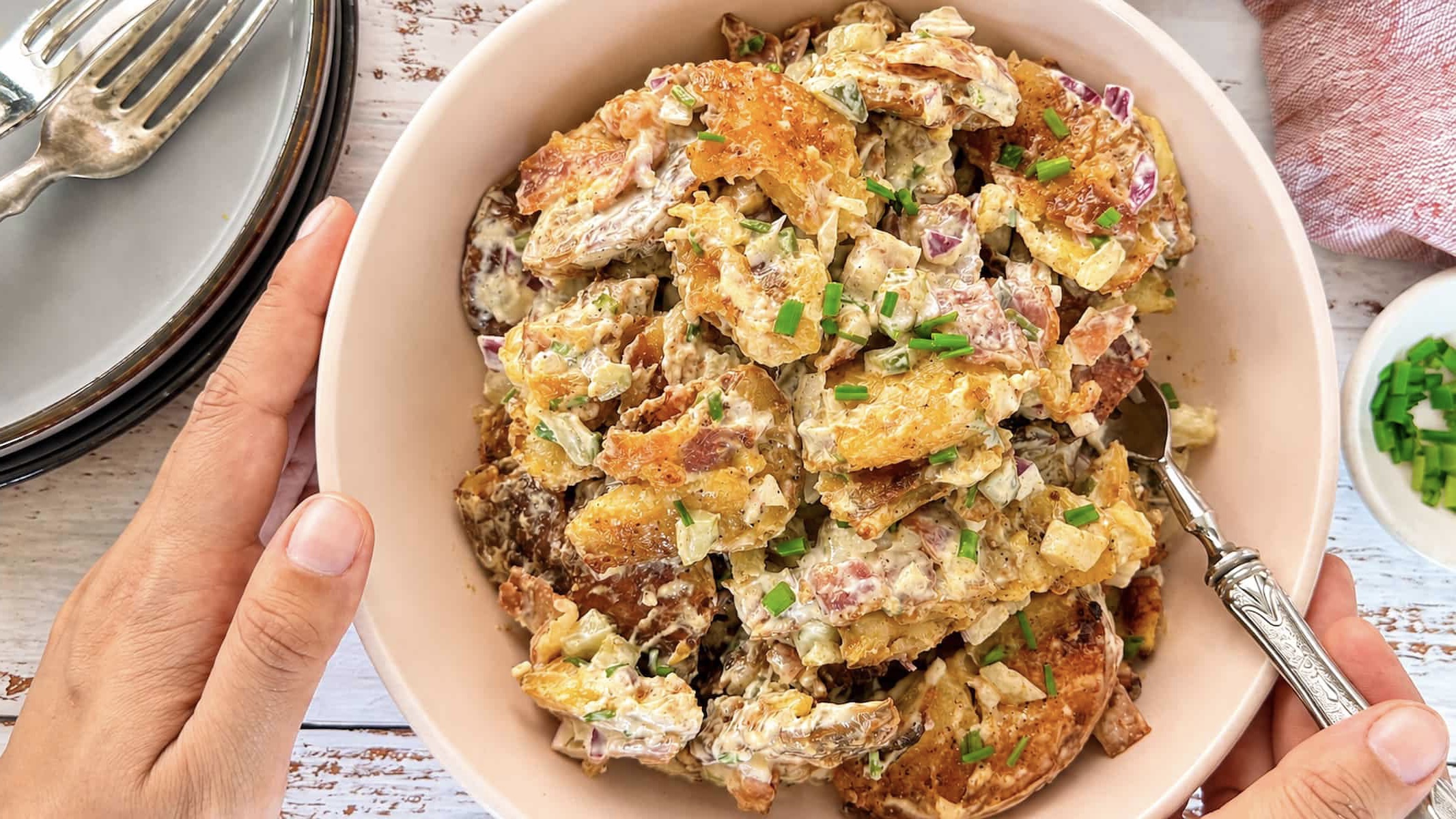 hand holding a bowl of smashed potato salad with crispy bacon, pickles, red onion, and fresh chives in a creamy dressing.