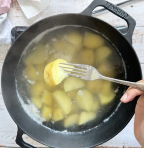 hand with a fork piercing a cubed potato in a water-filled pot with more.