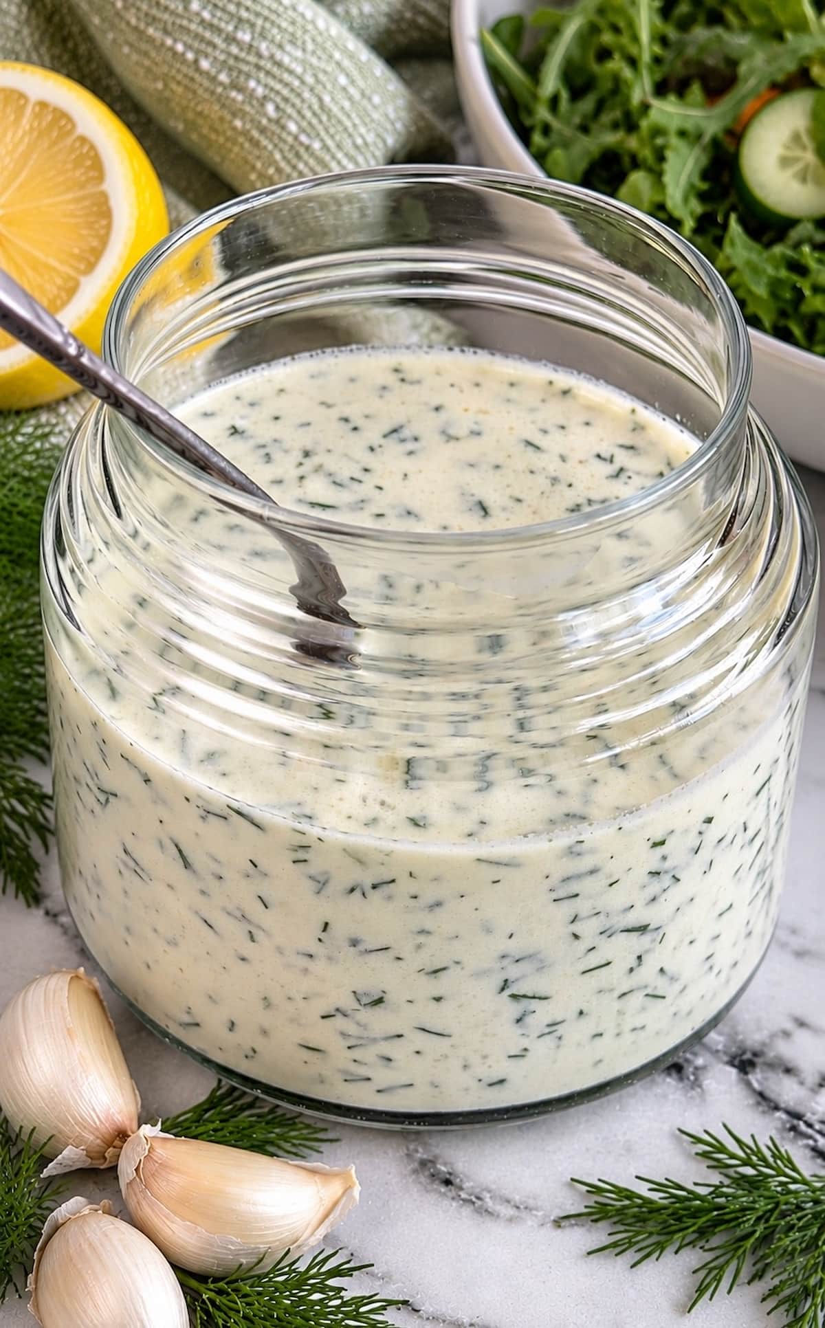 Creamy dill yogurt salad dressing in a glass jar with fresh dill, garlic, lemon, and a bowl of salad greens in the background.