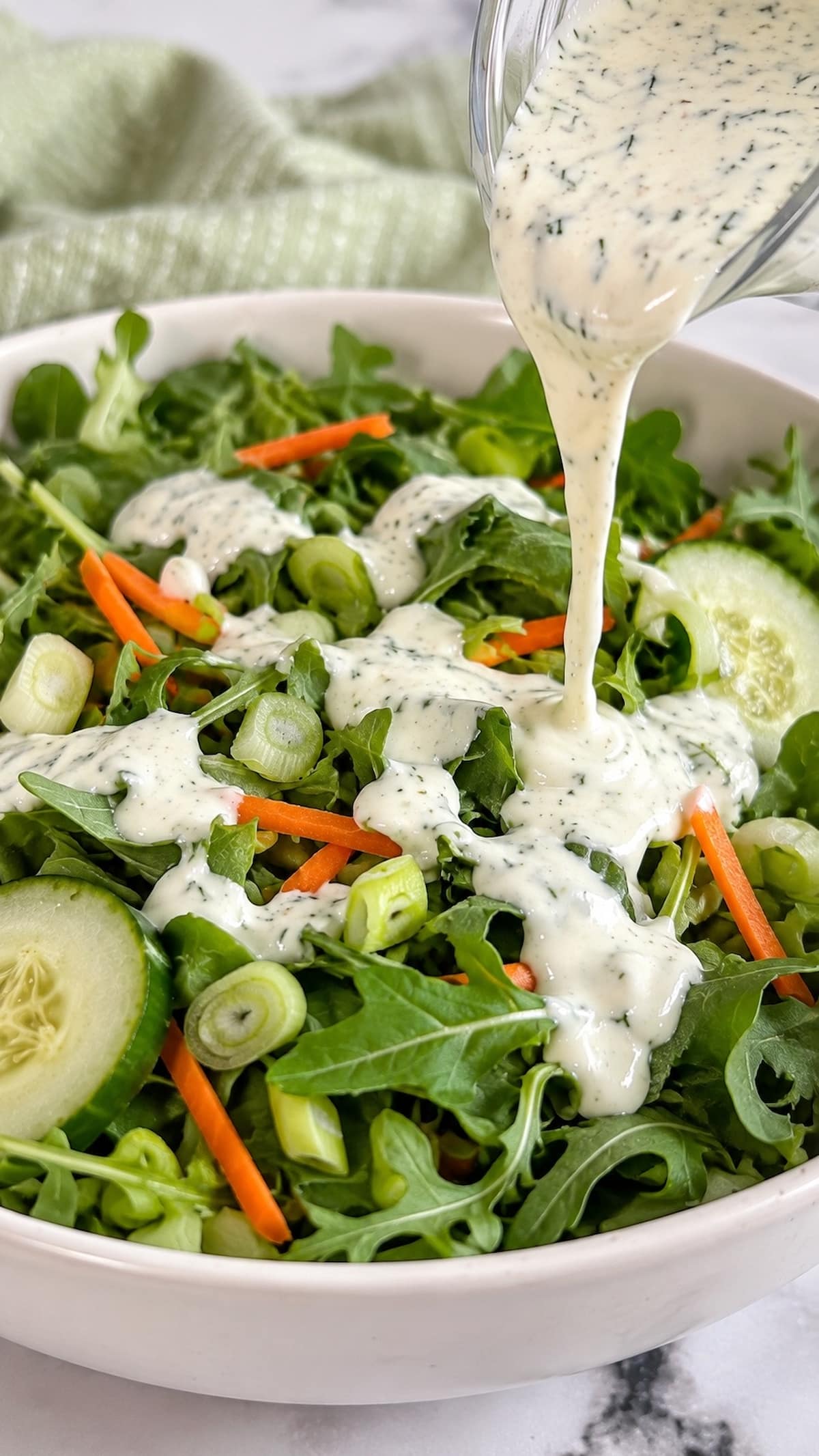 Creamy dill yogurt salad dressing being drizzled over fresh greens with cucumber, carrots, and green onions.