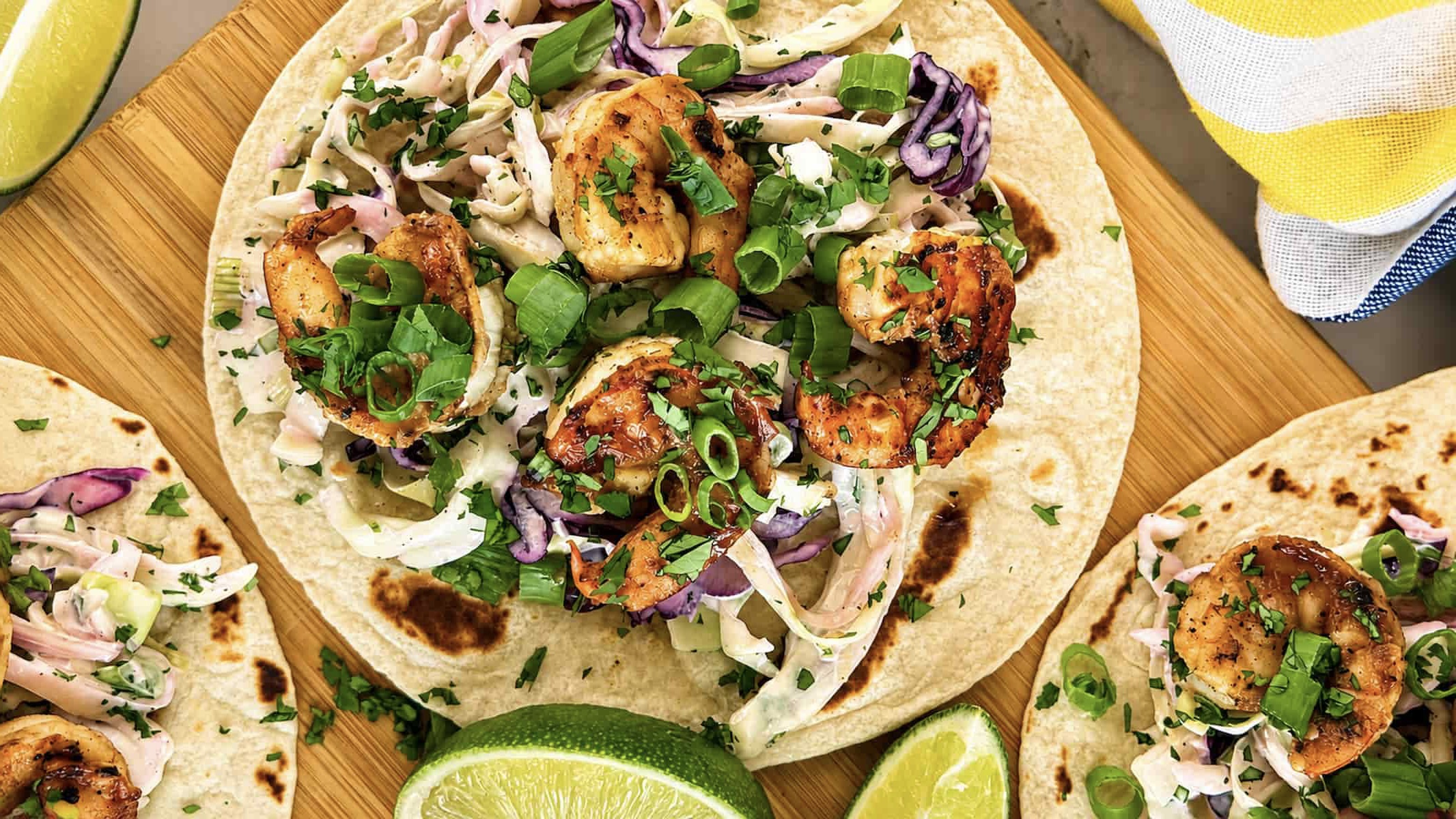 Jerk shrimp tacos with charred seasoned shrimp, creamy purple and green cabbage slaw, fresh cilantro, green onions, and lime wedges on warm flour tortillas.