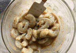 raw shrimp marinating in jerk sauce in a small glass bowl with a rubber spatula.