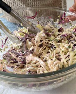 creamy red and green shredded cabbage coleslaw in a large glass bowl with tongs.