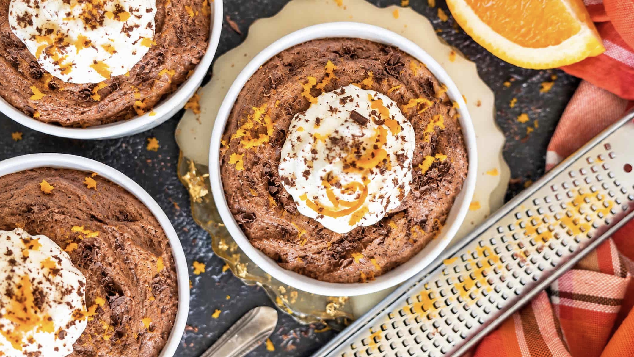 3 ramekins of chocolate orange mousse topped with whipped cream, fresh orange zest, and chocolate shavings next to an orange-zested microplane grater.